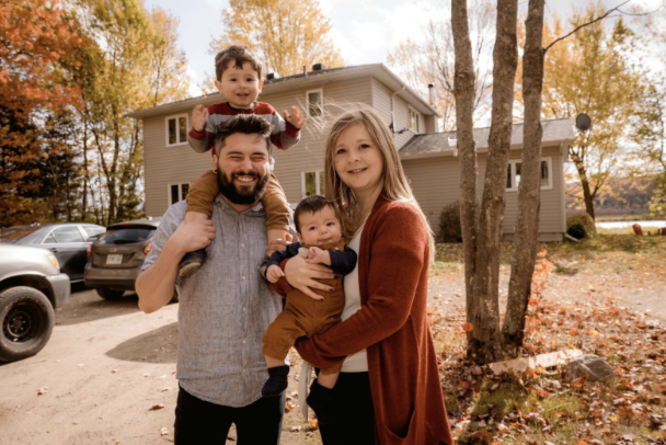 Smiling family of four standing outdoors in front of their home during autumn, with colorful trees and fallen leaves in the background. The father holds one child on his shoulders while the mother holds the younger child.